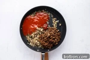 A frying pan on a white surface contains chopped onions and mushrooms, a portion of tomato sauce, and a serving of cooked lentils, all separated into sections and not yet mixed together.