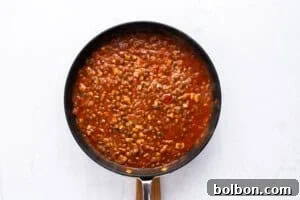 A black pan filled with a chunky red tomato sauce containing corn, lentils, and other visible vegetables, set against a white background.