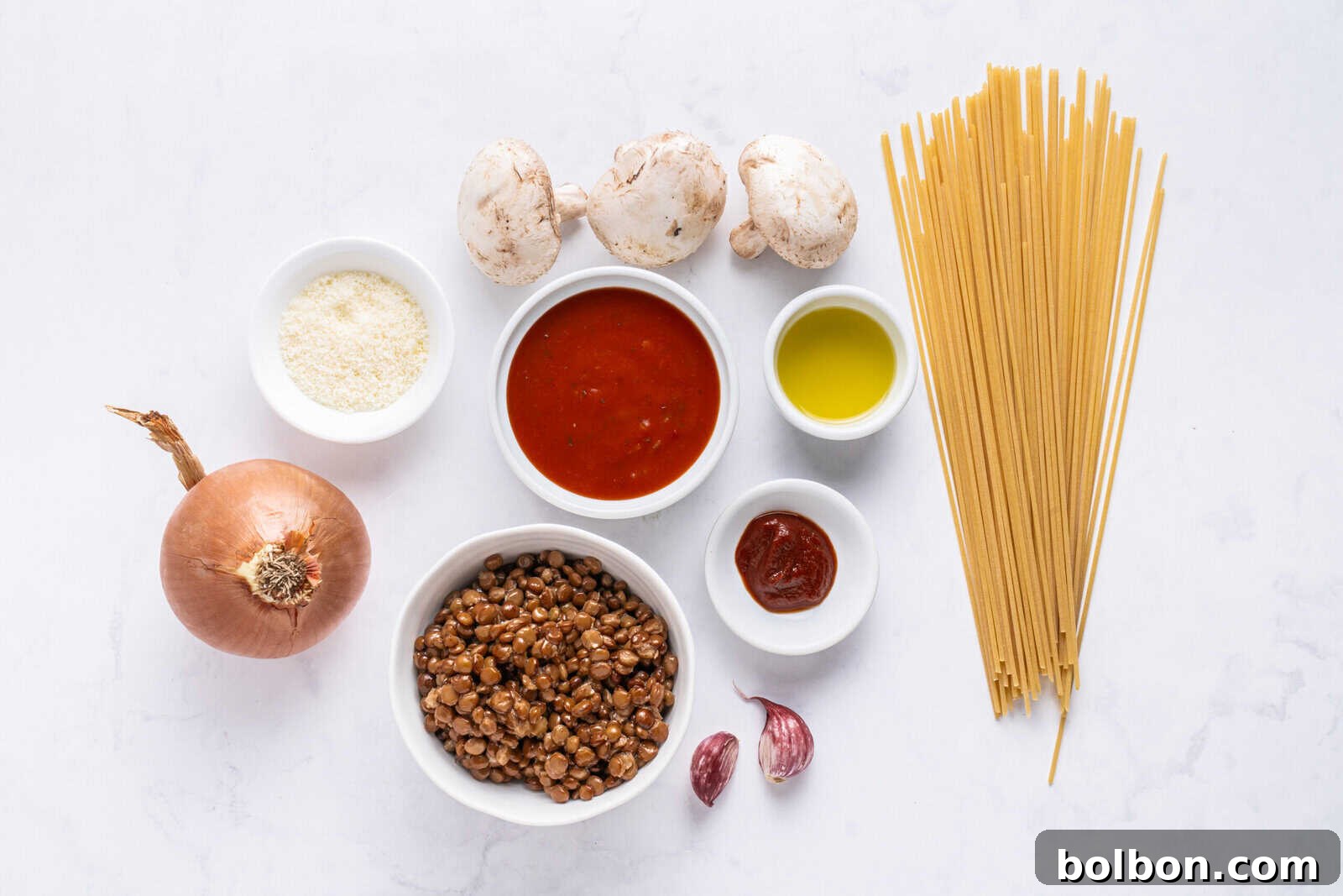 Ingredients for a pasta dish arranged on a white surface: uncooked spaghetti, whole mushrooms, yellow onion, lentils, tomato sauce, olive oil, tomato paste, grated cheese, and two garlic cloves in small bowls.