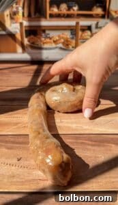 Golden Rice Paper Apple Delights 8 A hand with pale pink nail polish arranges a long, coiled sausage and a round sausage on a wooden table in a bakery, with shelves of bread and pastries blurred in the background.