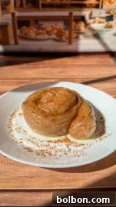 Golden Rice Paper Apple Delights 9 A cinnamon roll dusted with cinnamon sits on a white plate atop a wooden table, with a bakery display of various pastries blurred in the background.