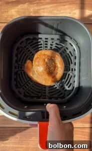 Golden Rice Paper Apple Delights 10 A hand is holding an air fryer basket containing a single coiled raw sausage, seasoned with spices, on a black grill tray. The background is a wooden surface illuminated by sunlight.