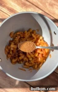 Golden Rice Paper Apple Delights 5 A spoonful of ground spices is held above a pot containing sautéed chopped onions and vegetables, ready to be added to the mixture. Warm sunlight casts shadows on the wooden surface beneath.