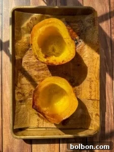 Two roasted acorn squash halves placed cut side up on a baking sheet lined with printed parchment paper, sitting on a wooden surface with sunlight casting shadows.