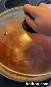 A hand holding the lid of a pot on a stovetop, with condensation visible on the glass lid and some food residue around the pot’s rim.
