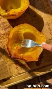 A hand uses a fork to scrape the inside of a roasted spaghetti squash half on a parchment-lined baking sheet.