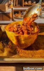 A halved roasted squash filled with a chunky meat and tomato sauce. A spoon scoops some of the filling, and the dish sits on a browned baking tray in sunlight, with bread and kitchen items blurred in the background.