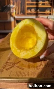 A hand holds half of a yellow melon with its seeds removed, revealing the hollow inside. Sunlight highlights the fruit’s juicy texture, and shelves with bread are visible in the background.