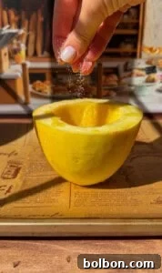 A hand sprinkles salt onto a halved spaghetti squash resting on a baking sheet, with a blurred background of a bakery display and bread.