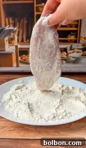 Mama's Italian Chicken Cutlets 8 A hand dips a raw chicken breast fillet into a plate of flour on a wooden table, preparing it for cooking. In the background, there are breads and other kitchen items.
