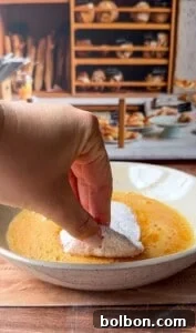 Mama's Italian Chicken Cutlets 9 A hand dips a powdered, crescent-shaped pastry into a bowl of beaten eggs, with shelves of bread and pastries blurred in the background.