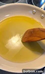 Mama's Italian Chicken Cutlets 11 A wooden spatula stirs oil in a light-colored frying pan on a stovetop, with light reflecting off the surface of the oil.