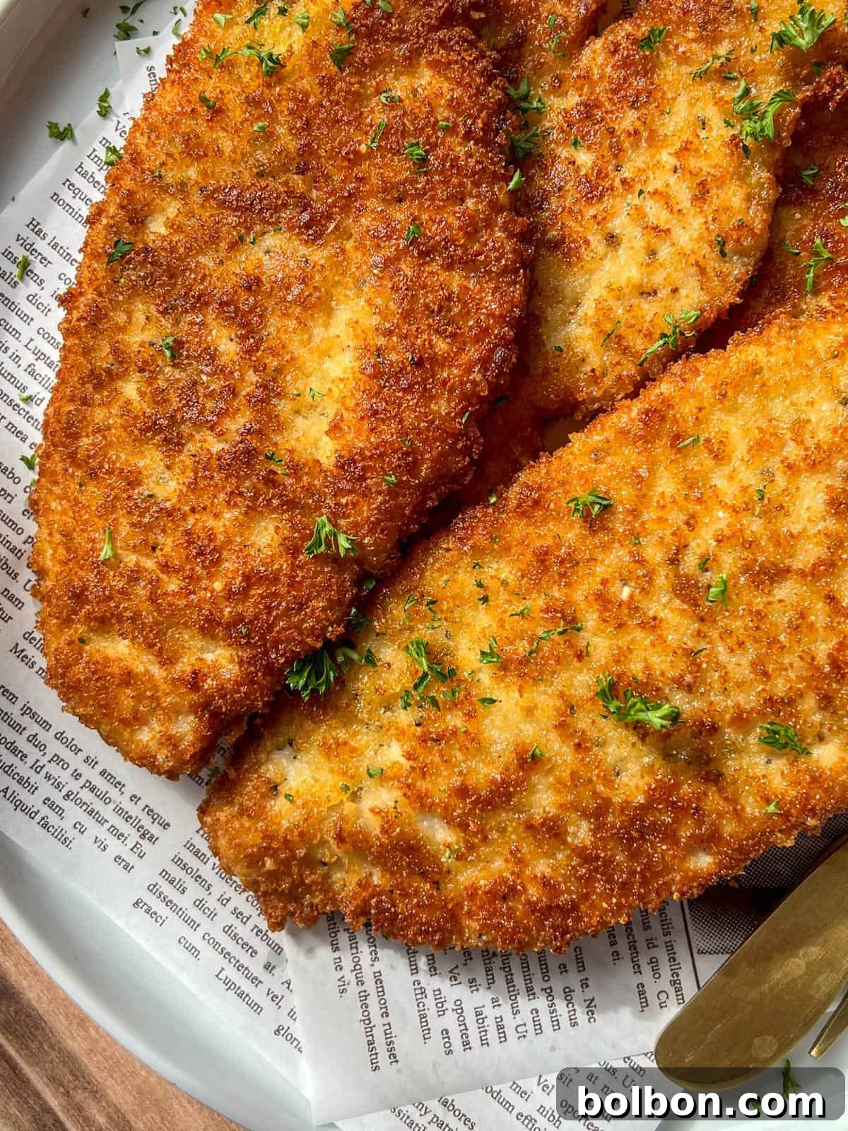 Mama's Italian Chicken Cutlets 3 Golden, crispy breaded chicken cutlets garnished with chopped parsley are served on a plate lined with printed newspaper-style parchment paper. A knife is partially visible at the edge of the plate.