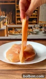 Mama's Italian Chicken Cutlets 5 A hand lifts a raw, thin slice of chicken breast from a white plate with more raw chicken, set on a wooden table in front of a bakery-themed background with bread and pastries.