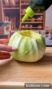 A hand holds a whole cabbage on a wooden cutting board while olive oil is poured over it from a green bottle; shelves with bread are visible in the blurred background.