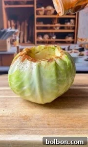 A whole green cabbage with the top cut off sits on a wooden surface. Seasoning is being sprinkled onto the open top, and there are shelves with bread and kitchen items blurred in the background.
