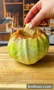A hand places a pat of butter into a hollowed-out green cabbage sprinkled with spices, set on a wooden cutting board with a kitchen scene blurred in the background.