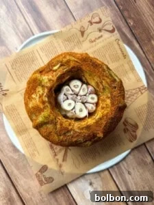 A round loaf of bread with a hollow center filled with halved garlic bulbs, placed on brown parchment paper atop a white plate on a wooden surface.