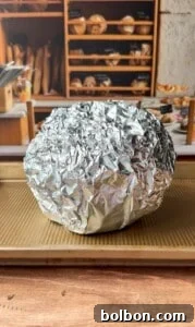 A round object completely wrapped in crinkled aluminum foil sits on a baking sheet with a bakery display of bread loaves in the background.