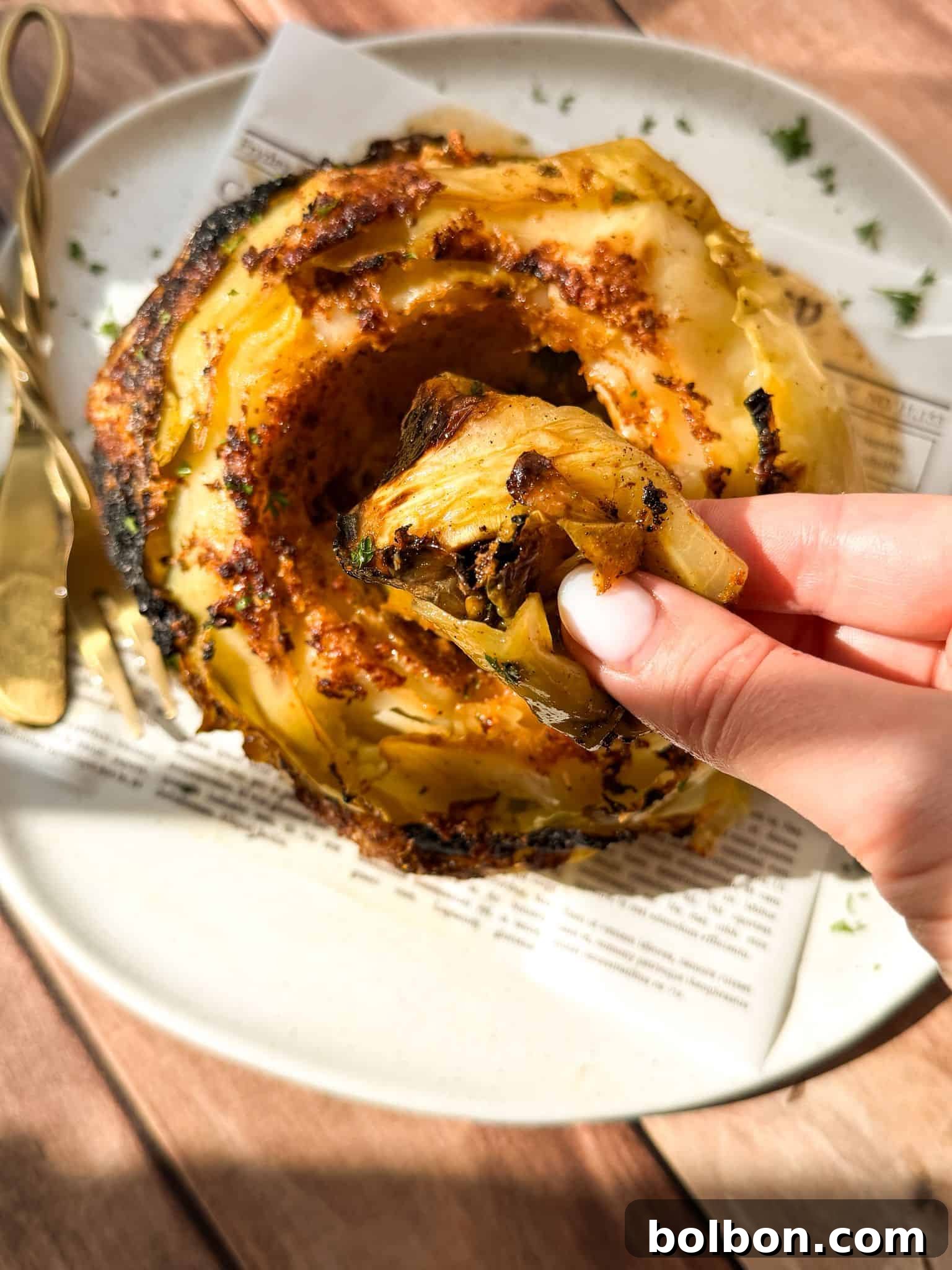 A hand holds a roasted cabbage leaf above a plate with a whole, charred roasted cabbage, garnished with herbs. A fork and knife rest on the plate, and sunlight highlights the food.