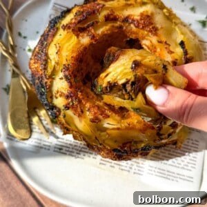A hand pulls a crispy roasted slice from a whole roasted cabbage served on a white plate with golden cutlery and a piece of printed paper underneath. Sunlight highlights the charred edges and seasoning.