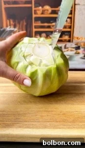 A hand holds a green cabbage on a wooden cutting board while a knife is inserted into the stem, preparing to cut it. Shelves with kitchen items are visible in the background.