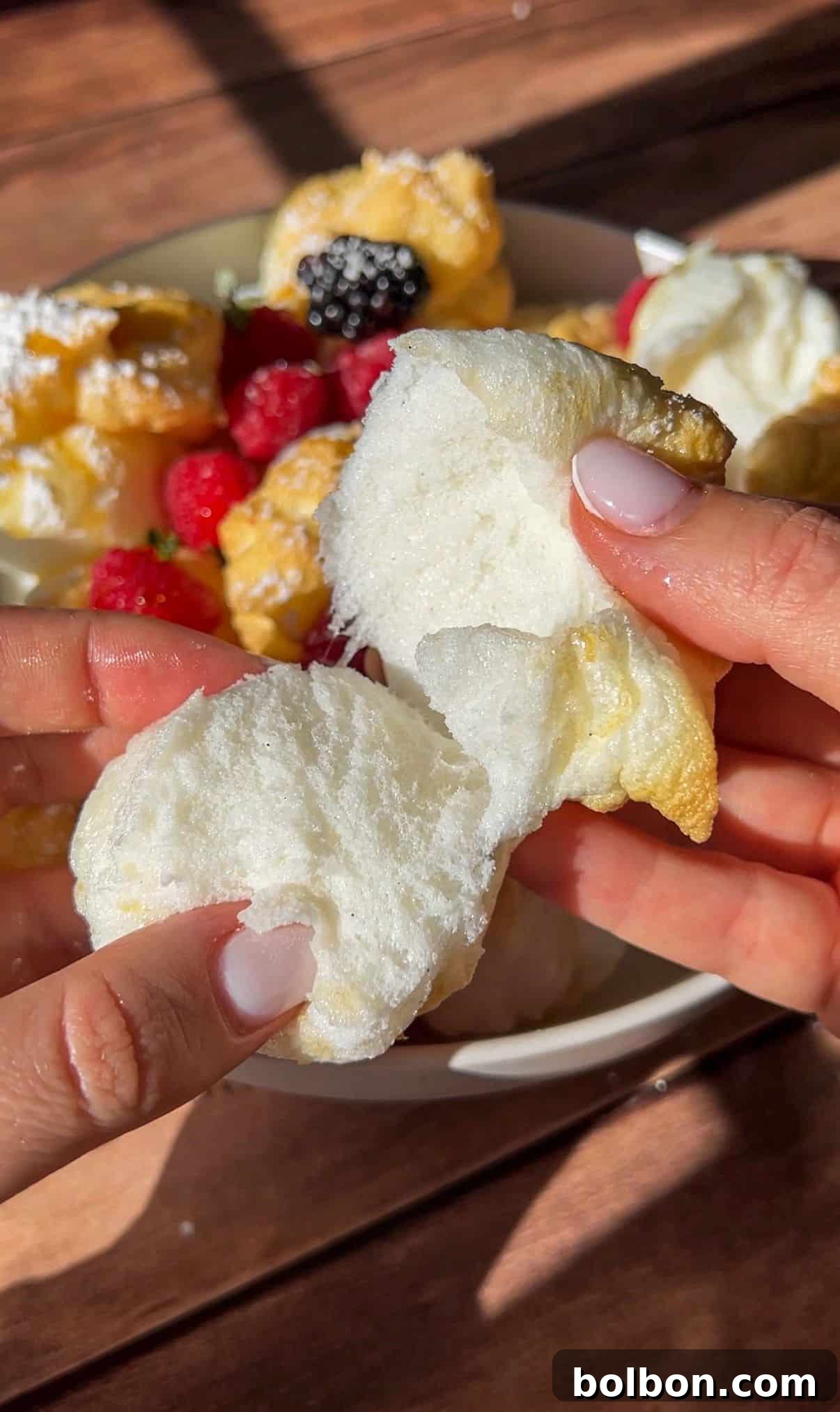 Protein Packed Pancake Bites 2 A person with light pink nails tears open a fluffy, white meringue cookie. In the background, a plate holds more meringues, High Protein Mini Pancakes (Viral), raspberries, blackberries, and powdered sugar, touched by sunlight.