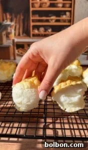 Protein Packed Pancake Bites 9 A hand with light-pink nails lifts a fluffy, golden-topped baked meringue from a cooling rack, with more meringues and shelves of bread in the blurred background.