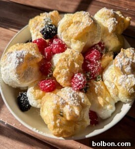 Protein Packed Pancake Bites 10 A bowl of golden, fluffy cream puffs dusted with powdered sugar, garnished with fresh raspberries, blackberries, and sprigs of thyme, all bathed in warm sunlight on a wooden surface.