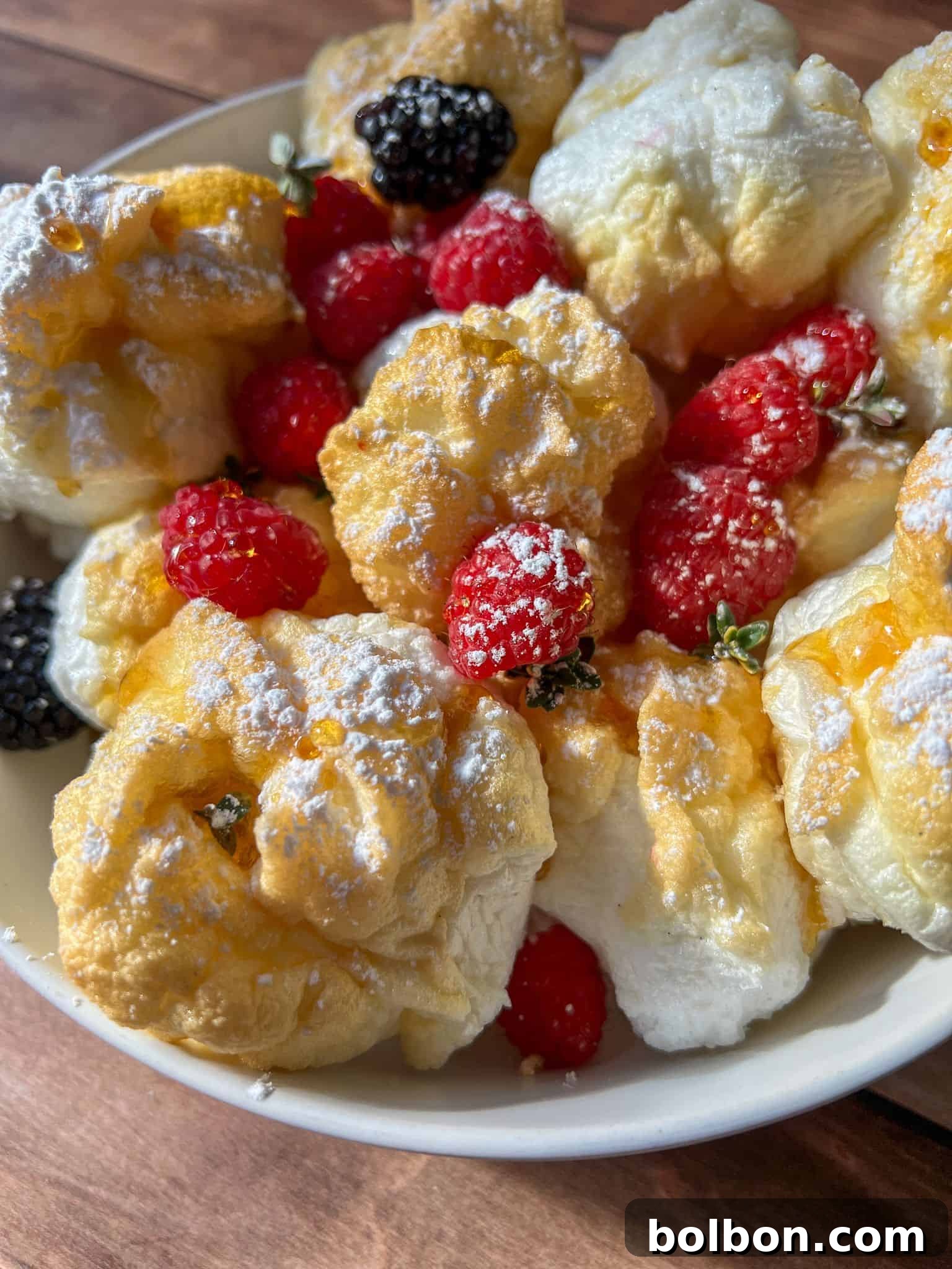 Protein Packed Pancake Bites 3 A bowl of fluffy cream puffs dusted with powdered sugar, garnished with fresh raspberries and blackberries, sits beside High Protein Mini Pancakes (Viral) on a rustic wooden table.
