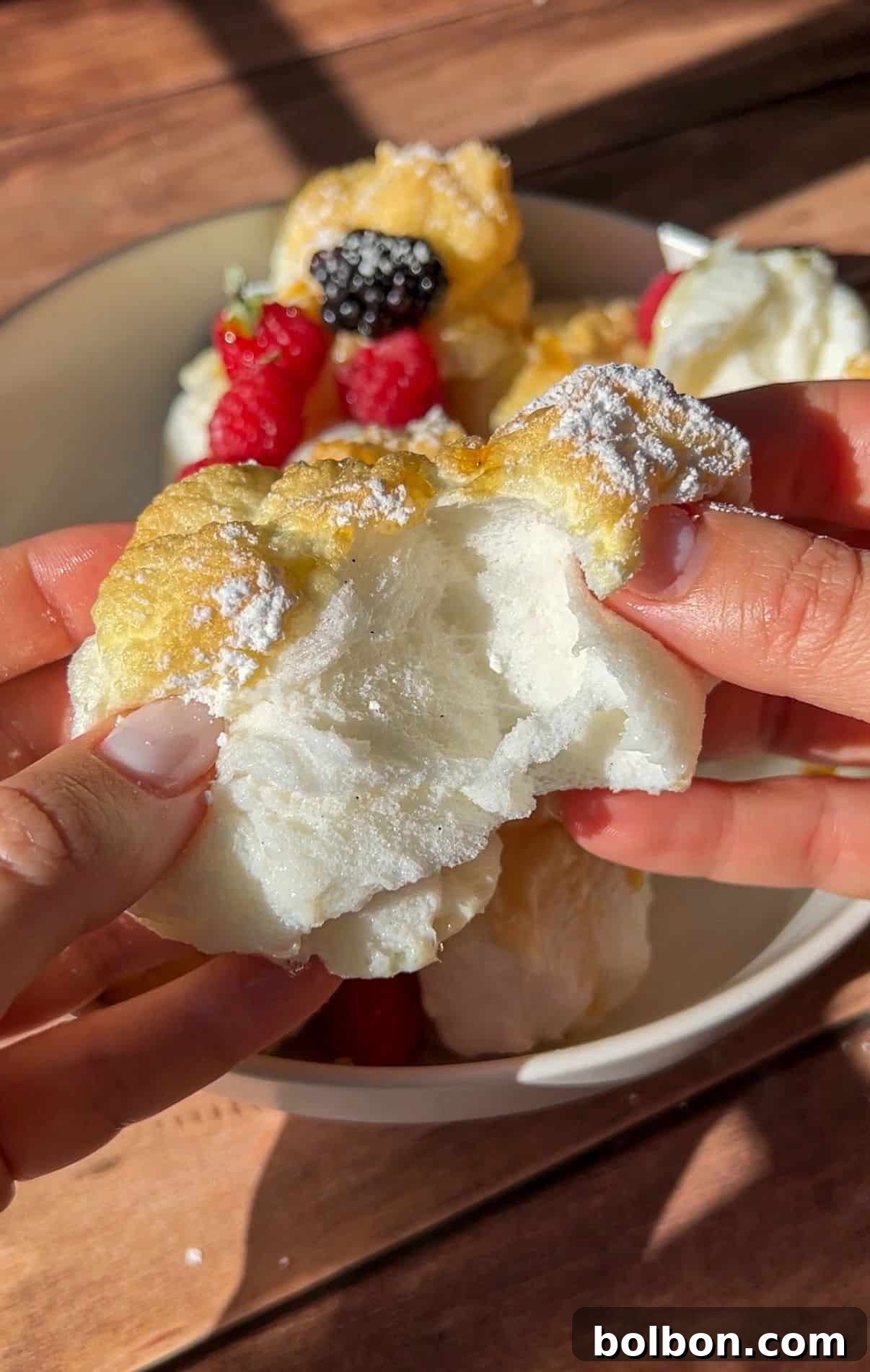 Protein Packed Pancake Bites 4 A person breaks open fluffy, golden High Protein Mini Pancakes (Viral) dusted with powdered sugar. Fresh berries are visible on other pieces of the pancakes in a white bowl on a wooden table.