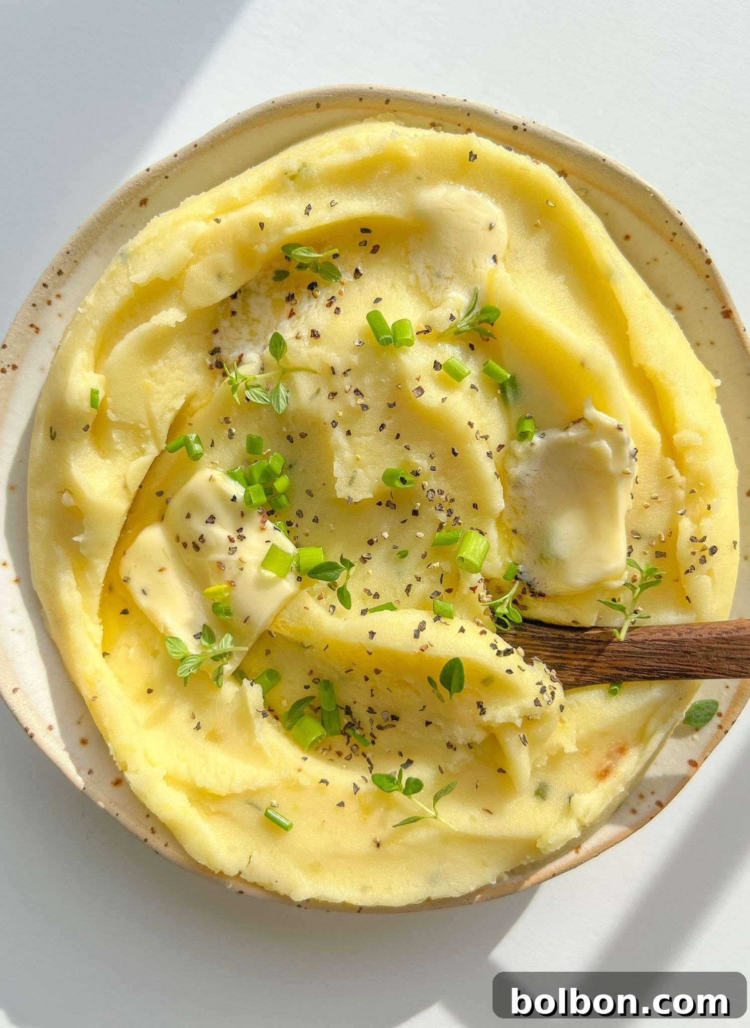 Golden Garlic Mash 3 A plate of creamy Roasted Garlic Mashed Potatoes topped with melted butter, cracked black pepper, and fresh green herbs, with a wooden spoon resting invitingly in the dish.