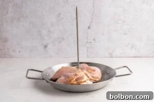 Aloha Chicken Gyro 6 Raw, marinated chicken pieces are placed in a round metal pan with handles, and a vertical metal skewer stands upright through the center of the chicken. The background is a light, textured surface.