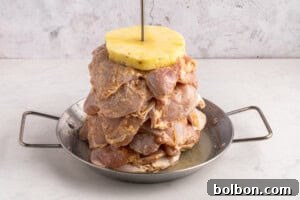 Aloha Chicken Gyro 8 Raw marinated meat stacked on a vertical skewer with a slice of pineapple on top, placed in a metal pan with handles, against a light textured background.