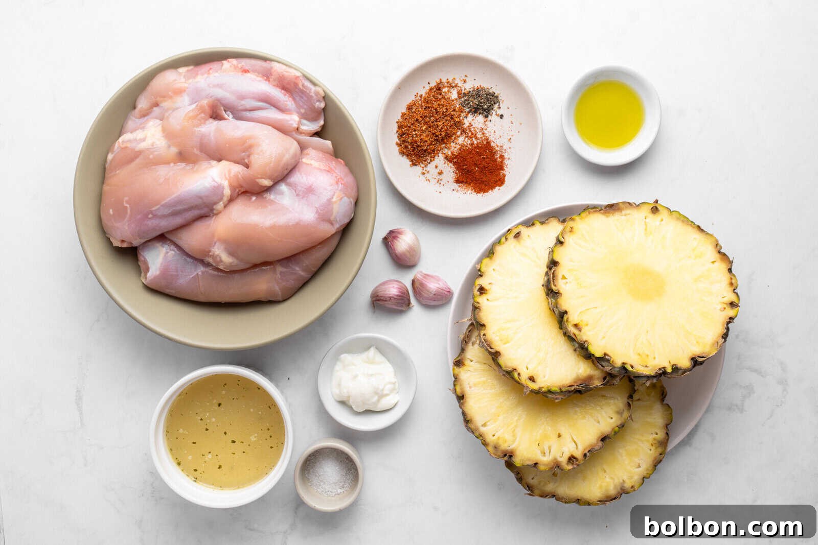Aloha Chicken Gyro 3 Flat lay of raw chicken thighs, sliced fresh pineapple, garlic cloves, assorted spices, olive oil, a bowl of marinade, a small dish of mayonnaise, and a dish of salt on a white surface.