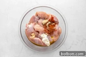Aloha Chicken Gyro 4 Raw chicken pieces in a glass bowl with dollops of yogurt, minced garlic, oil, and red spices on top, ready to be mixed, on a white marble surface.