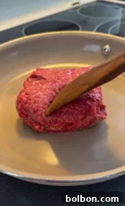 Layered Cabbage Comfort 6 A wooden spatula presses down on a chunk of raw ground beef in a beige frying pan on a stovetop.