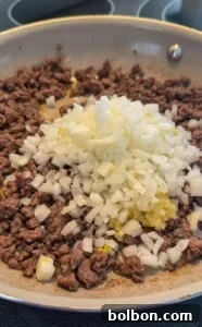 Layered Cabbage Comfort 8 Ground beef is browning in a skillet with a pile of freshly chopped onions and minced garlic on top, ready to be mixed together and cooked further.