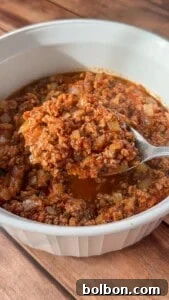 Layered Cabbage Comfort 10 A close-up of a spoonful of ground meat in a rich red sauce with diced onions, held over a white bowl filled with the same mixture, on a wooden surface.