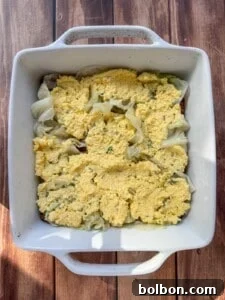 Layered Cabbage Comfort 12 A white square baking dish containing a layer of cooked cabbage topped with an uneven layer of yellow, crumbly cornbread mixture, sitting on a wooden surface.