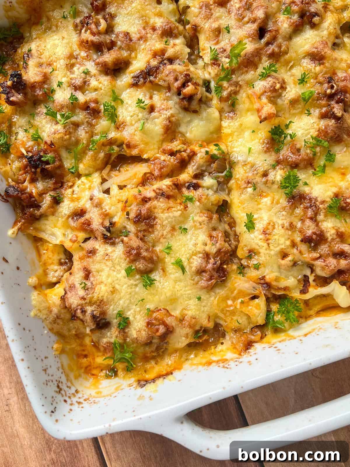Layered Cabbage Comfort 3 A close-up of a Cabbage Lasagna casserole in a white dish, topped with melted cheese, browned ground meat, and sprinkled with fresh chopped parsley on a wooden surface.