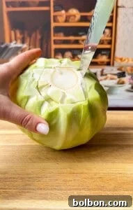 Layered Cabbage Comfort 4 A hand holds a green cabbage on a wooden cutting board while a knife cuts around its core. Shelves with bread and baked goods are visible in the blurred background.