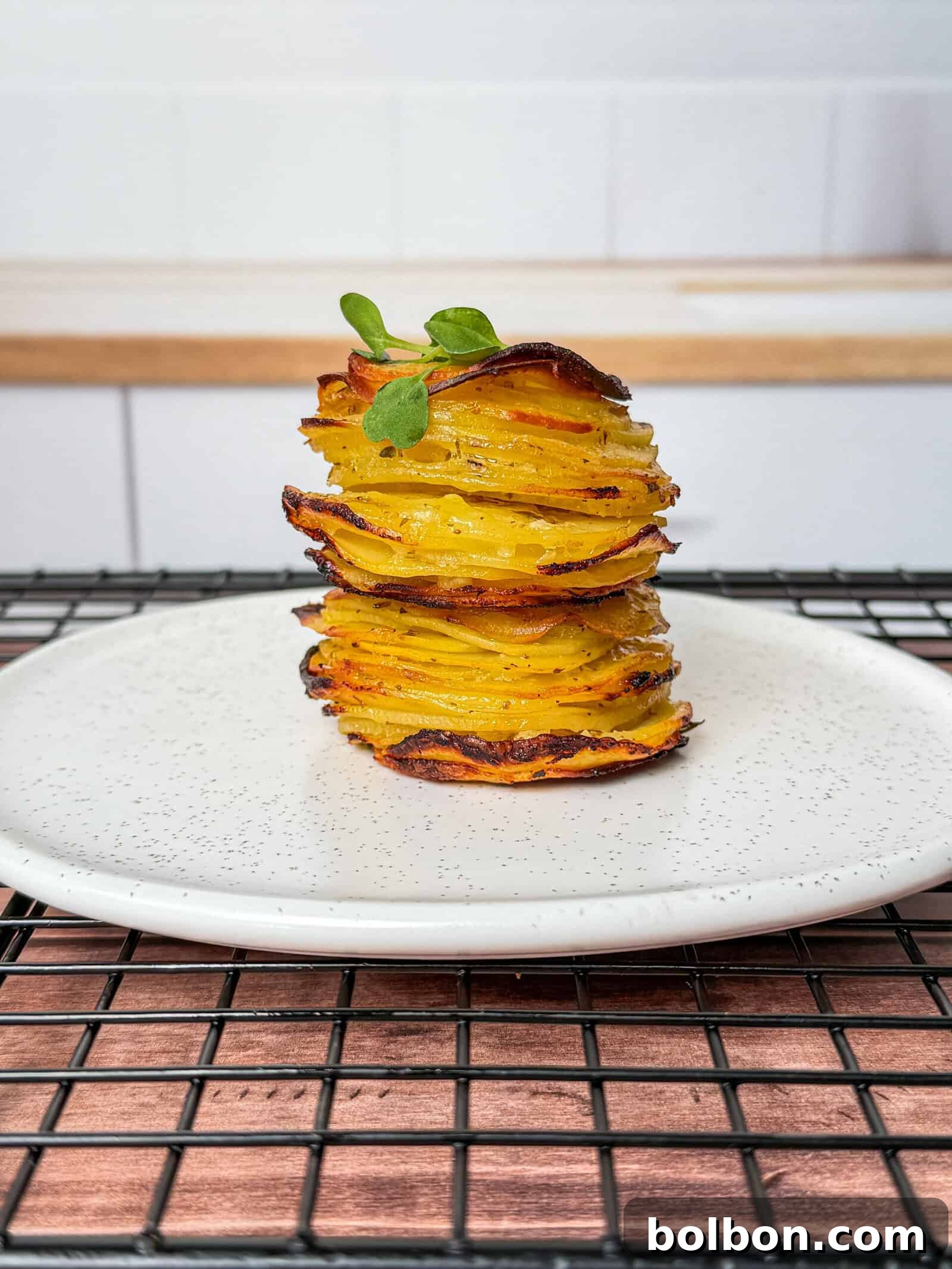 A neatly stacked tower of greek lemon potato stacks, thinly sliced and roasted, garnished with a small green herb, served on a white plate with a speckled pattern, placed on a black cooling rack.