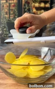 A hand uses a mandoline slicer to cut yellow potatoes into thin slices for Greek lemon potato stacks over a glass bowl on a wooden surface, with a festive, blurred background.