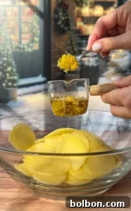 A hand pours olive oil and grated lemon zest from a measuring cup onto sliced potatoes in a glass bowl, preparing festive greek lemon potato stacks, with a wintery outdoor scene visible through the window in the background.