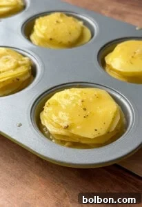 Close-up of a muffin tin filled with greek lemon potato stacks—thinly sliced potatoes seasoned with herbs and spices, ready to be baked. The tin rests on a wooden surface.