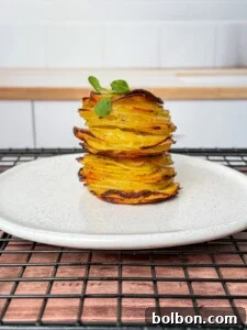A neatly stacked tower of greek lemon potato stacks, thinly sliced and roasted, garnished with a small green herb, served on a white plate with a speckled pattern, placed on a black cooling rack.