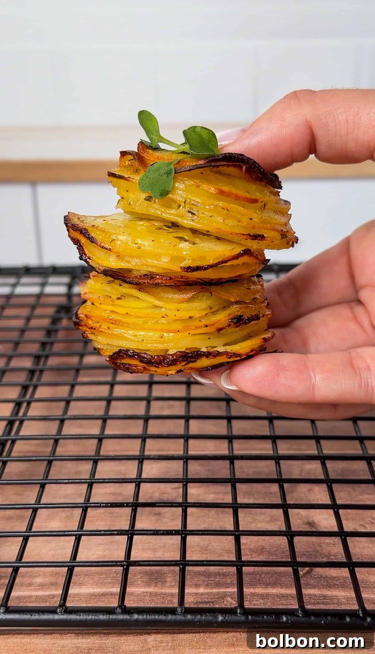 A hand holds a stack of crispy, golden greek lemon potato stacks, thinly sliced and garnished with a sprig of green herb, above a black cooling rack on a wooden surface.