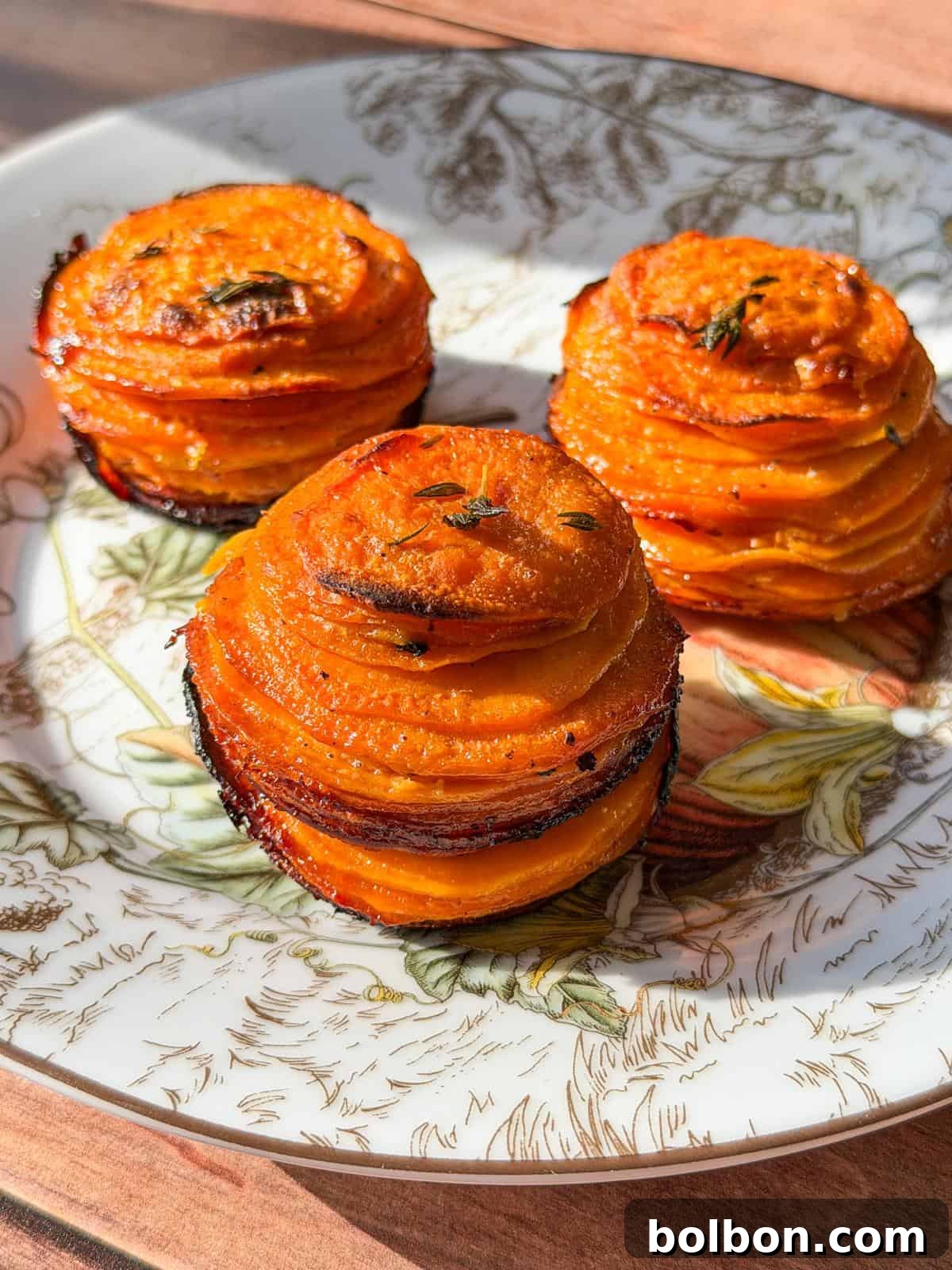 Three stacks of thinly sliced, roasted sweet potatoes, browned at the edges, are arranged on a decorative plate with a botanical pattern. Sunlight highlights their golden-orange color and crisp texture.