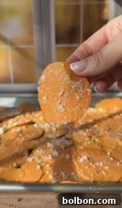 A hand holds up a round, orange sweet potato slice sprinkled with grated cheese, with more Sweet Potato Stacks and cheese spread out on a baking sheet in the background. Sunlight streams through a window behind.